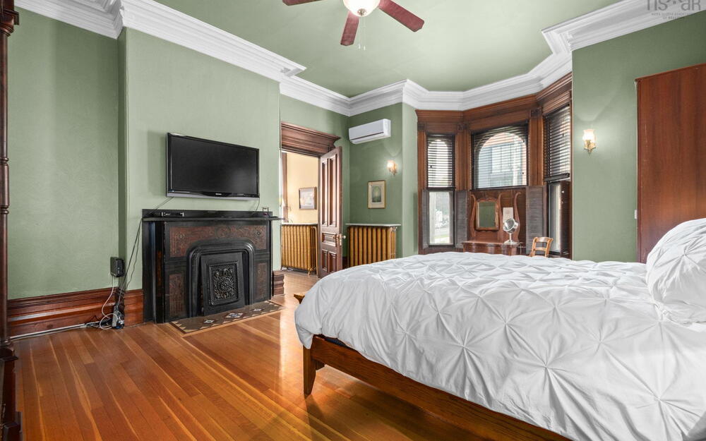Ornate fireplace mantel with decorative iron inlay, detailed crown molding, polished wood-finish flooring, deep-set bay windows with dark wood casings, and wall-mounted climate control unit