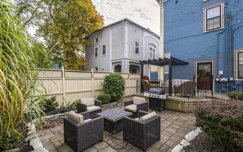 Stone patio featuring a raised deck, black pergola, privacy fencing, and mature landscaping